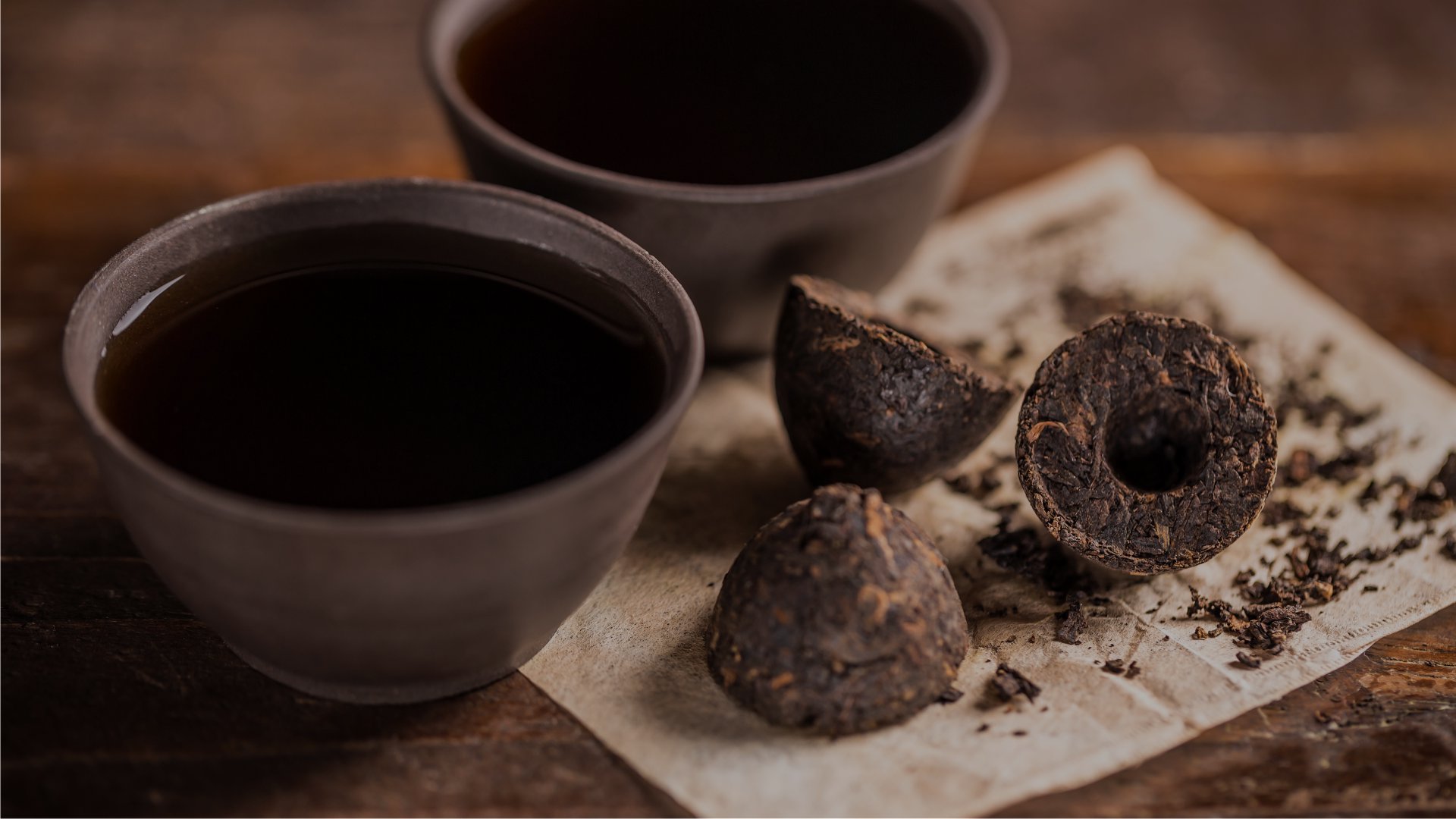 Dwie czarki z ciemną herbatą oraz sprasowane kulki herbaty pu-erh, z kawałkami rozkruszonymi na pergaminie, ustawione na drewnianym blacie.