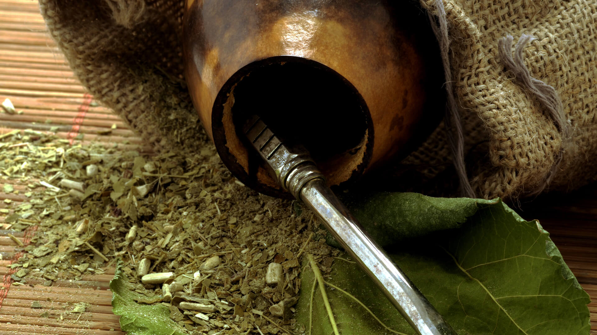 Tradycyjna tykwa z rurką do picia yerba mate, wysypane suszone liście i zioła, obok jutowy woreczek i świeże zielone liście na bambusowej macie.